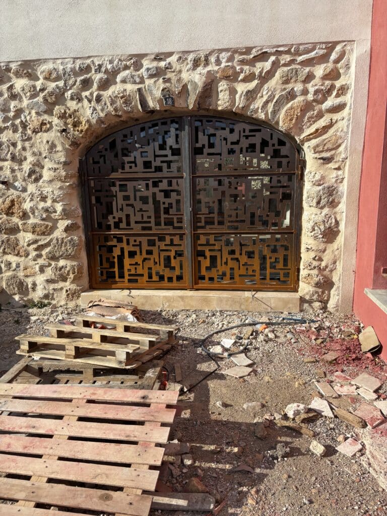 Portail métallique design à motifs géométriques noirs et rouille, mur en pierre n°18, chantier et palettes.